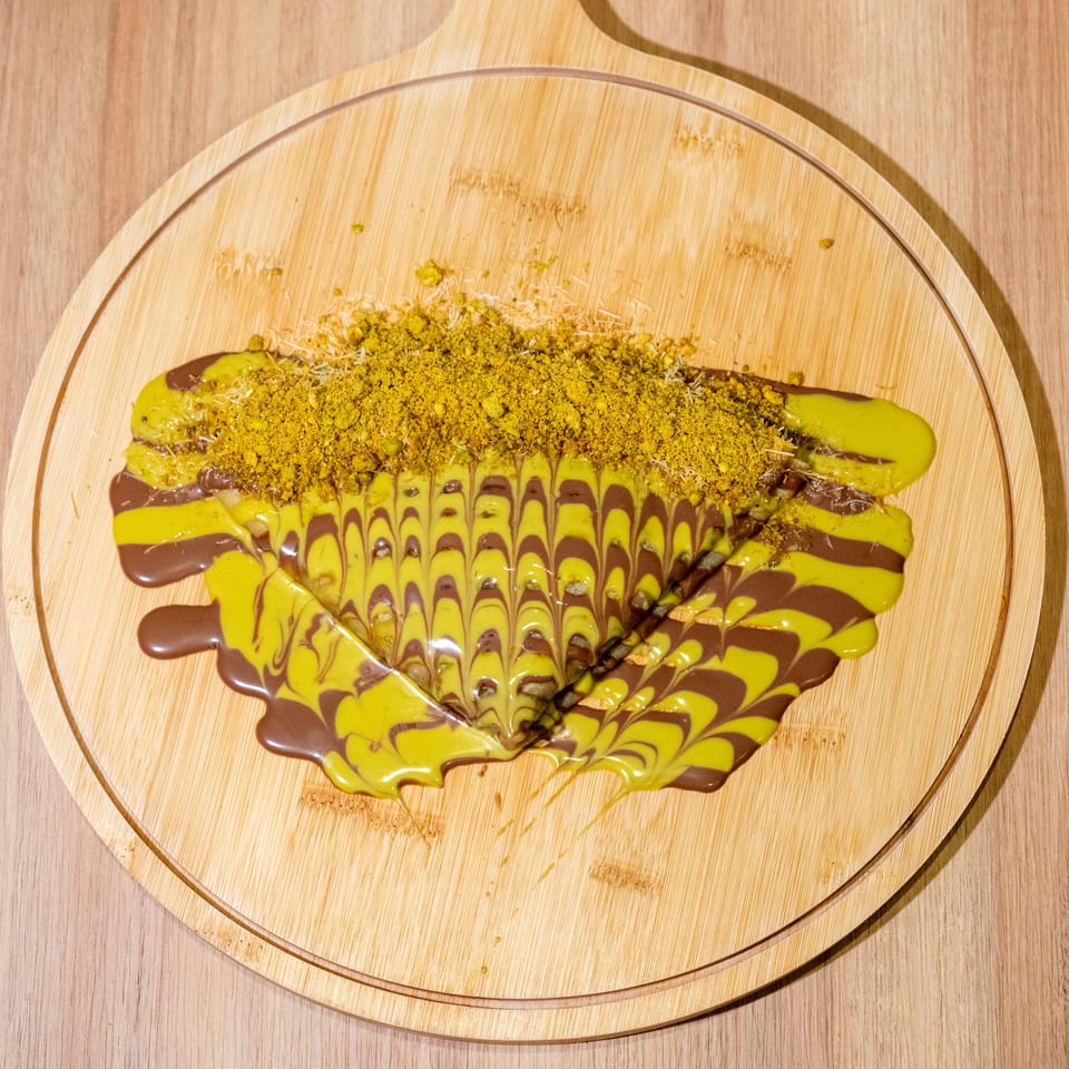 Triangular dessert drizzled with chocolate and yellow sauce, topped with crushed pistachios on a wooden serving board.
