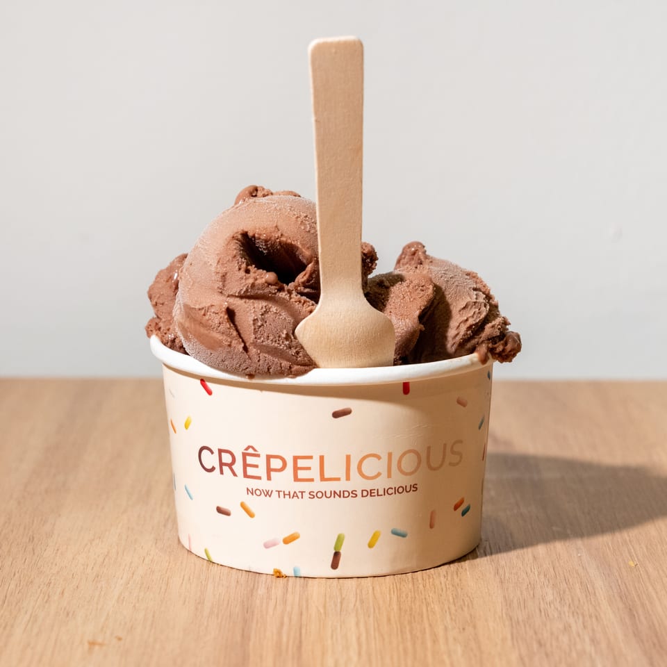Scoop of chocolate ice cream in a sprinkle-decorated Crêpelicious cup with a wooden spoon, on a wooden table.
