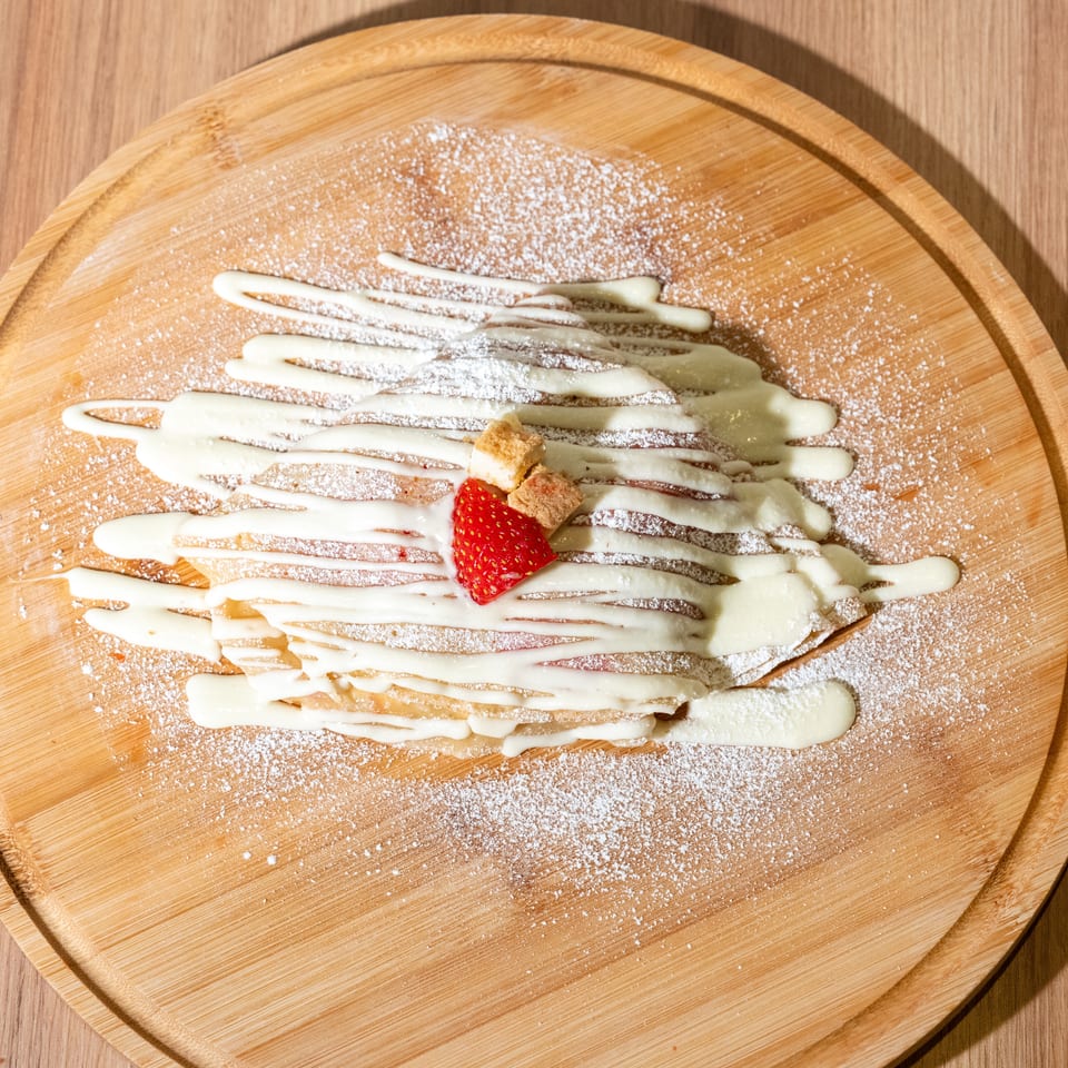 Dessert crepe drizzled with white sauce, dusted with powdered sugar, and topped with a strawberry and biscuit pieces on a wooden board.