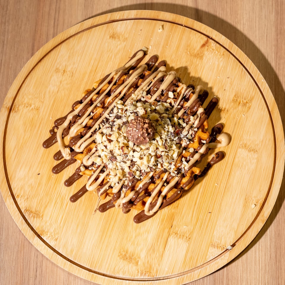 Waffle fries drizzled with chocolate and cream sauces, topped with chopped nuts and a chocolate-covered scoop on a wooden platter.