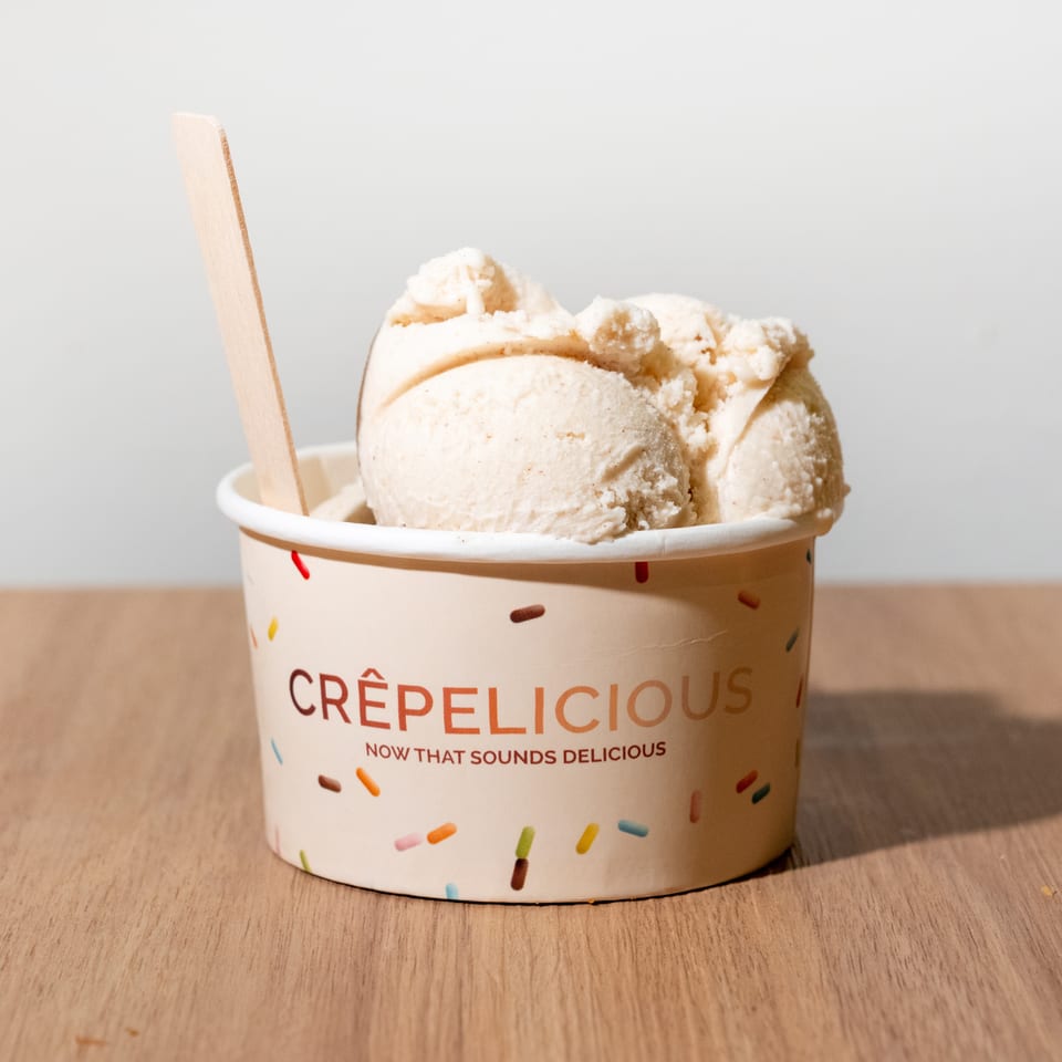 Several scoops of vanilla ice cream in a sprinkle-decorated Crêpelicious cup with a wooden spoon.