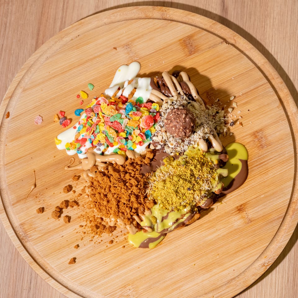 Overhead view of a heavily topped dessert on a round wooden board, covered with colorful cereal, chocolate, pistachio, crumbs, and sauces.