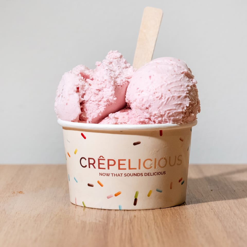 Three scoops of pink ice cream in a sprinkle-decorated Crêpeli'cious cup with a wooden spoon.